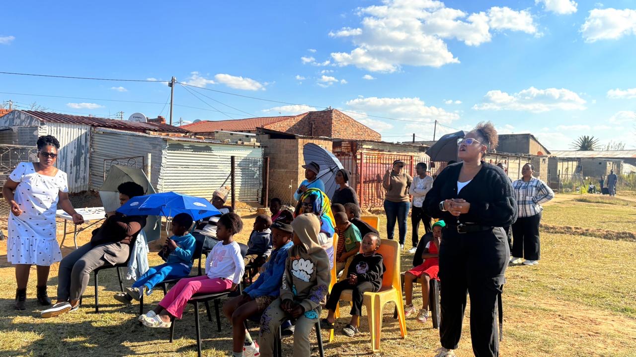Children and adults seated on chairs listening to a speaker outdoors