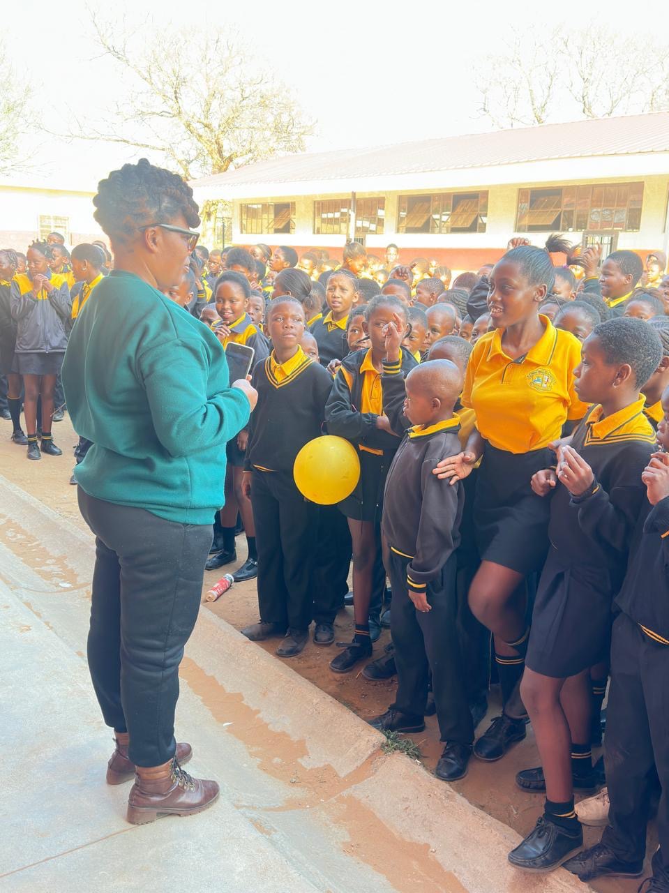 A volunteer addressing a large group of primary school learners outdoors